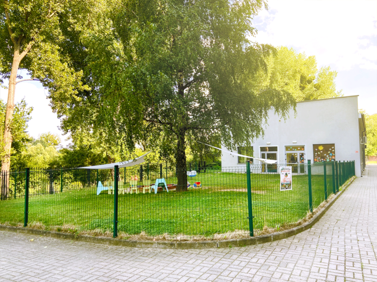 podwórko żłobka otoczone roślinami i drzewami, z widokiem na rzekę Oławka.