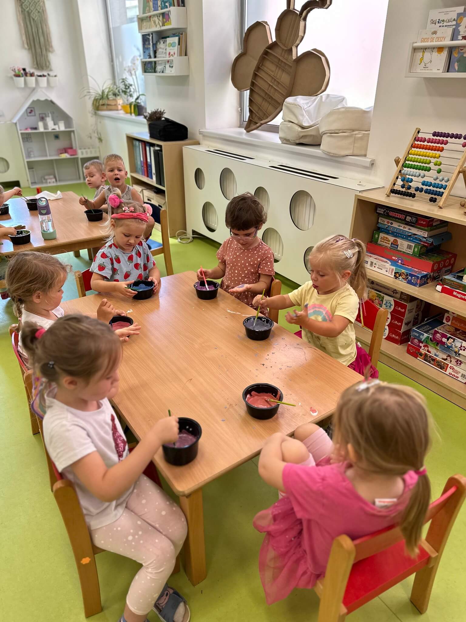 Grupa przedszkolaków siedzi przy stoliku i miesza kolorowe farby w miseczkach.