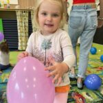 Mała dziewczynka trzyma w rączkach różowy balon i uśmiecha się do obiektywu.